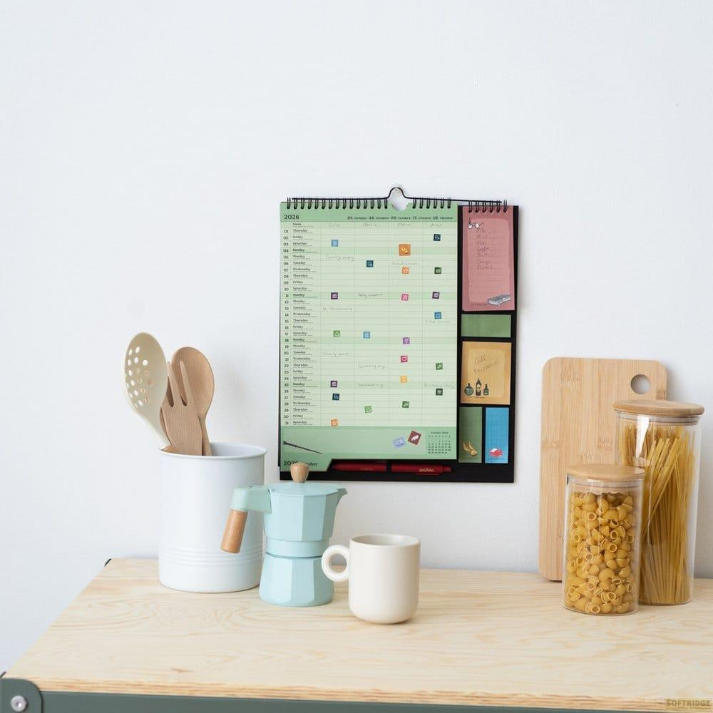 Calendario de pared, bote de utensilios, cafetera, taza y tarros de cristal con pasta en una mesa de madera.