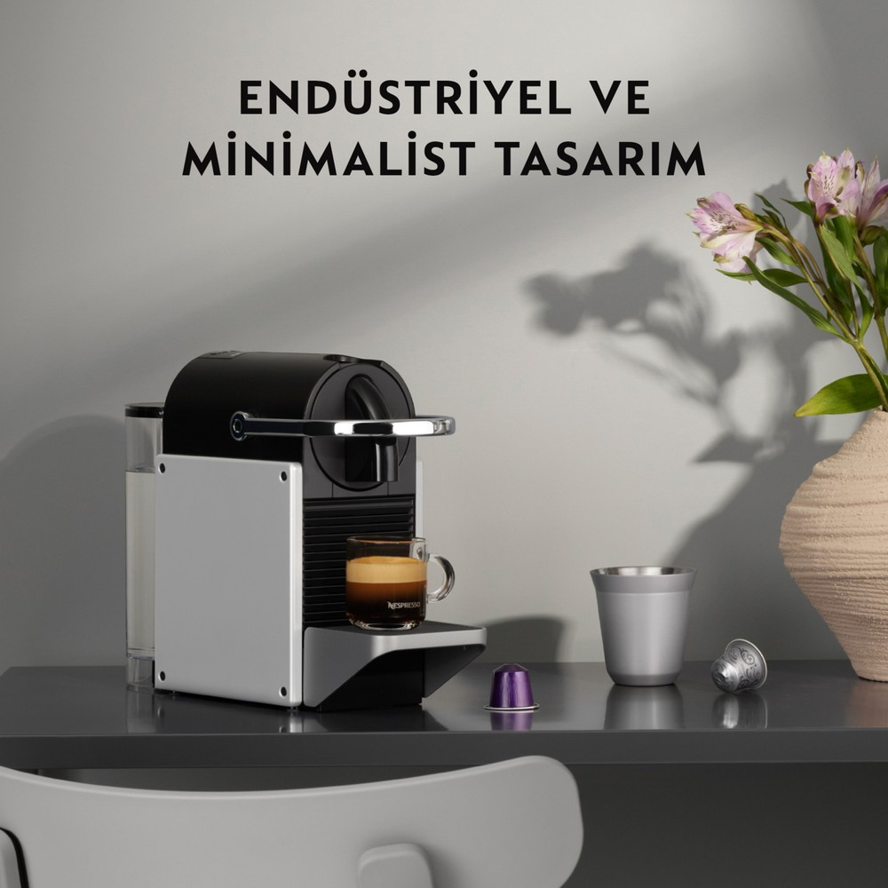 Siyah ve gümüş renkli kahve makinesi, pembe çiçekli vazo yanında koyu renk masada ve üstte yazı.