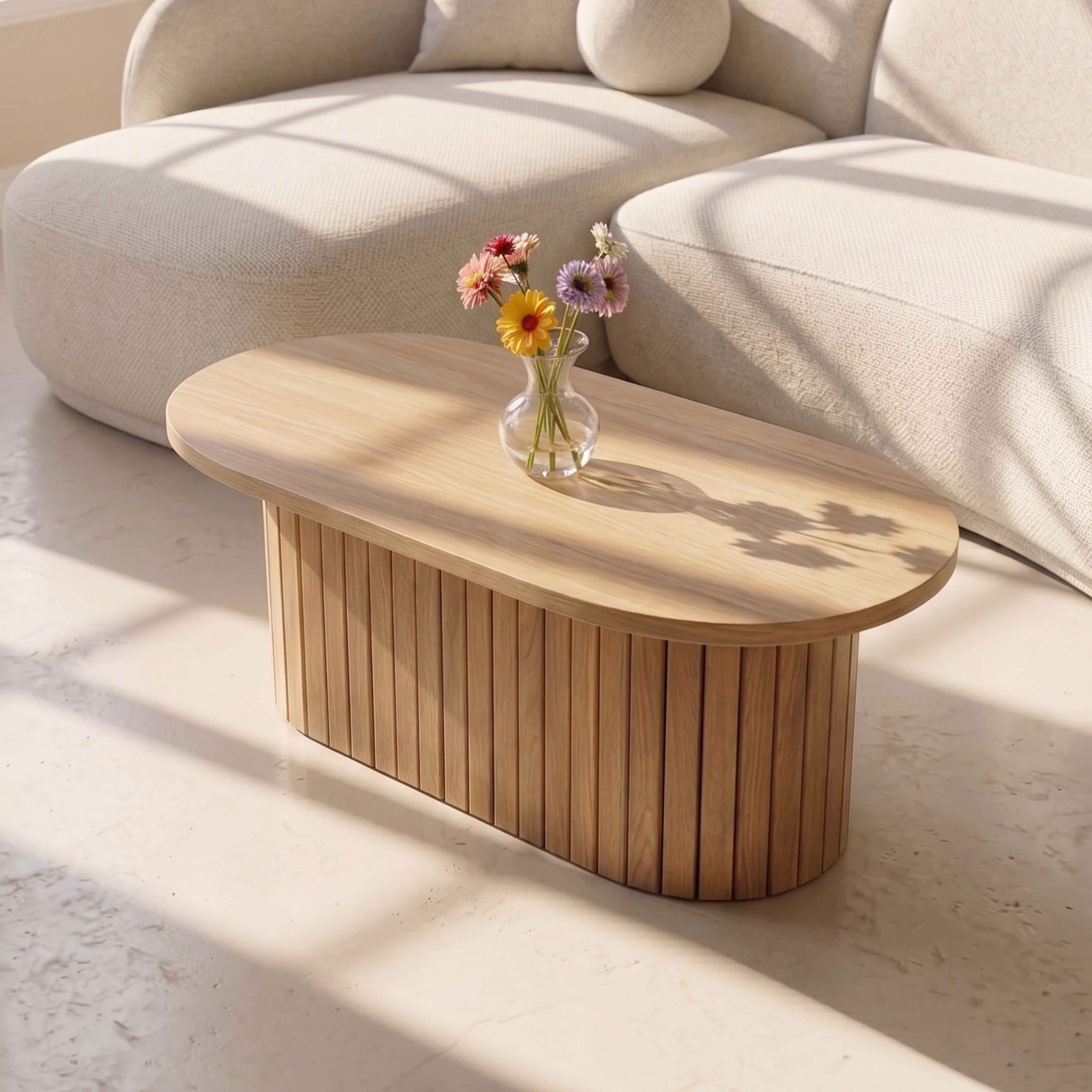 Ein ovaler Holztisch mit einer Blumenvase steht vor einem hellen Sofa in einem sonnigen Raum.