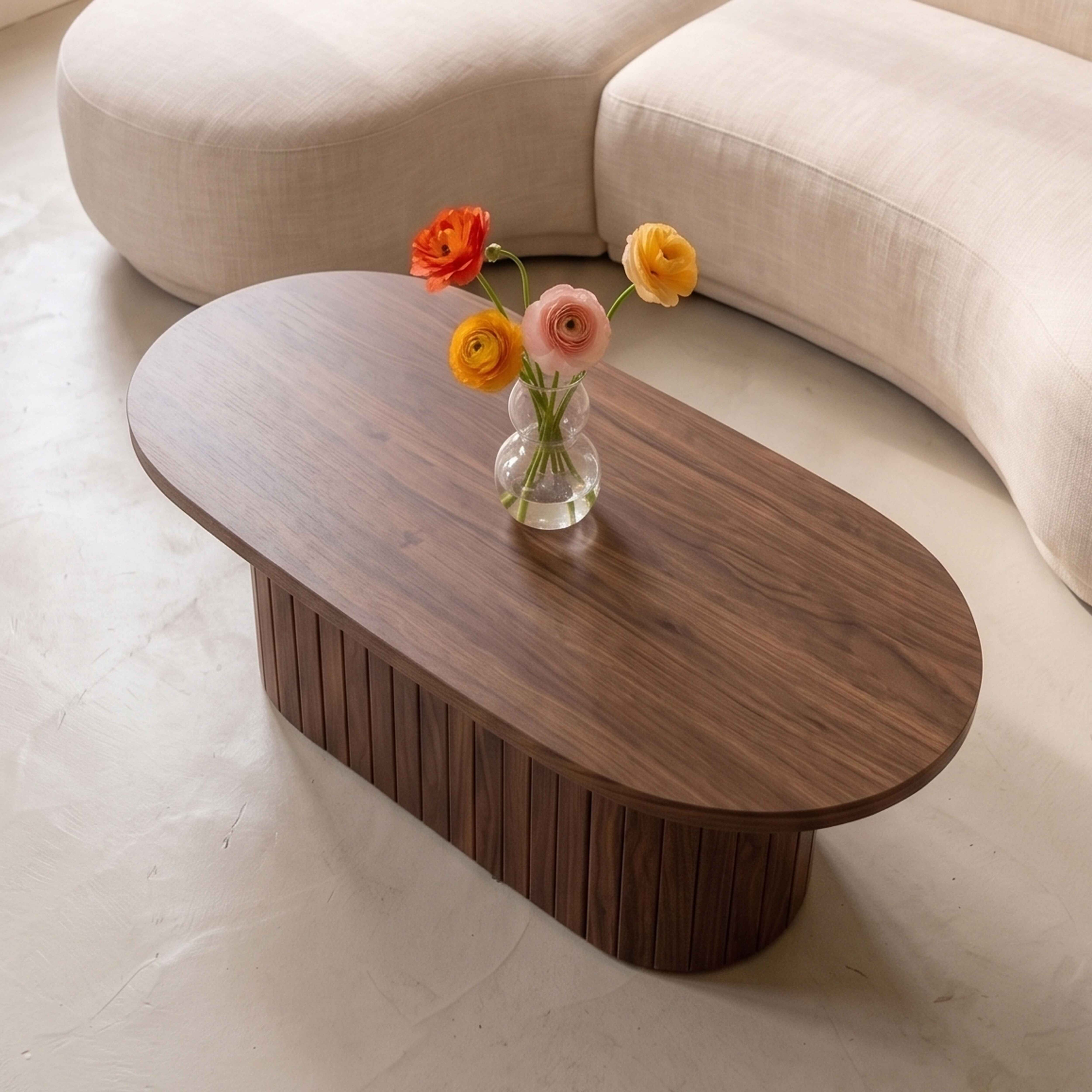 Ein ovaler Holzcouchtisch mit einer kleinen Glasvase voller bunter Blumen vor einem geschwungenen Cremesofa.