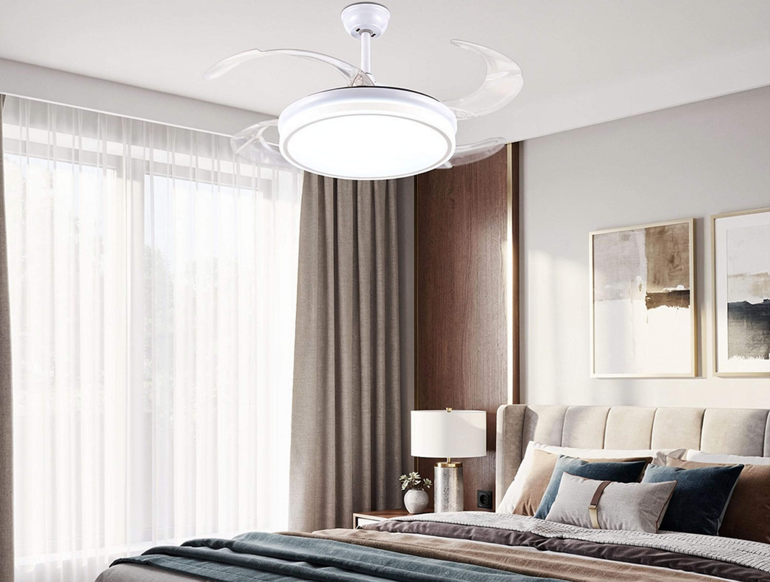Un dormitorio moderno con ventilador de techo iluminado, cama con cojines, cortinas y cuadros en la pared.