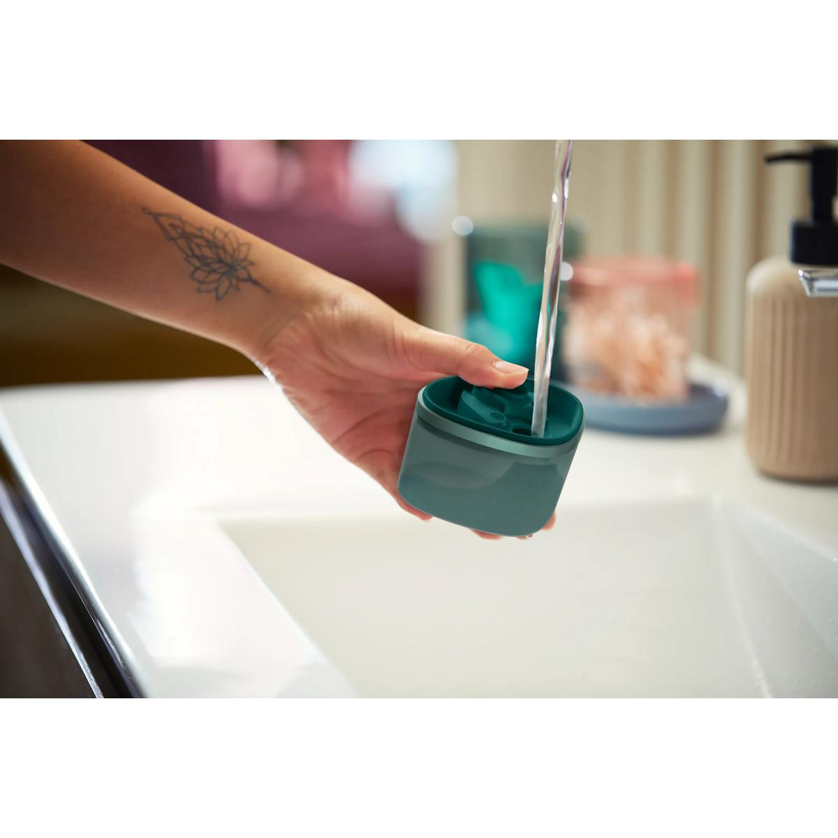 Una mano tatuata tiene un contenitore verde sotto l'acqua corrente di un lavandino.