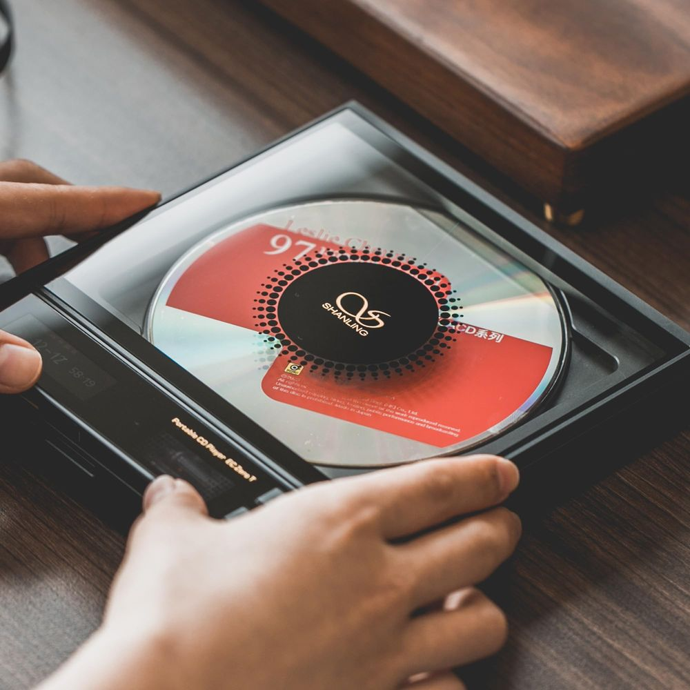 Manos sosteniendo un reproductor de CD portátil negro con un disco rojo y plateado en una mesa de madera.