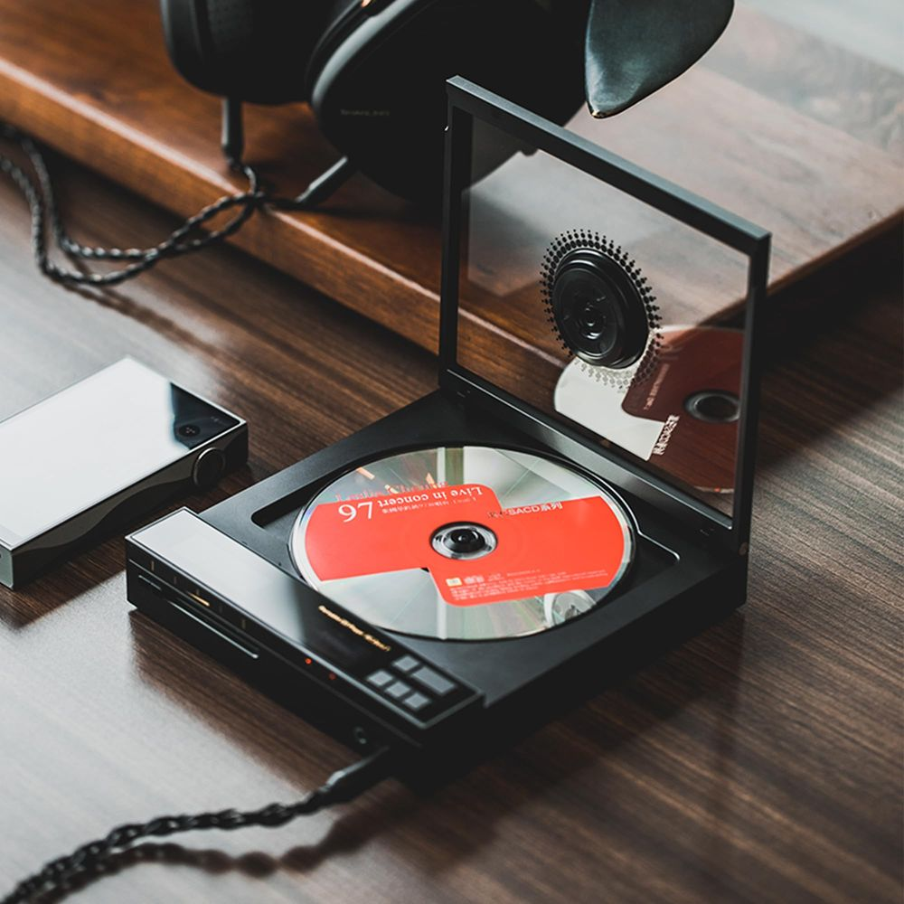 Un reproductor de CD portátil negro con tapa de vidrio abierta que muestra un disco rojo y plateado en un escritorio.
