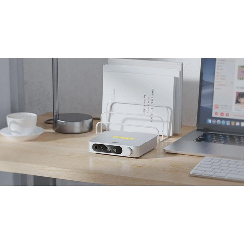 Un amplificador de audio plateado en un escritorio de madera junto a una laptop, taza y un organizador.