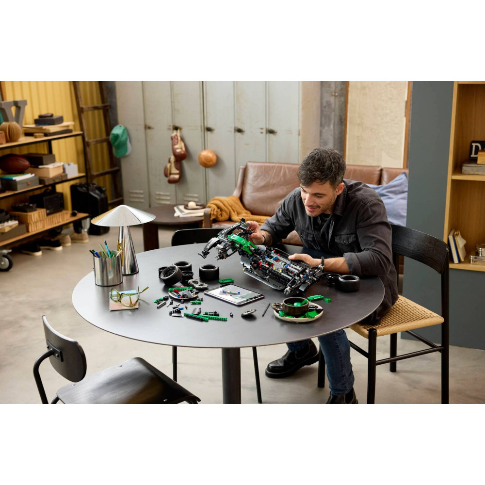 Yuvarlak bir masada oturan adam, etrafına saçılmış küçük parçalar ve bir tabletle bir Lego Technic araba monte ediyor.