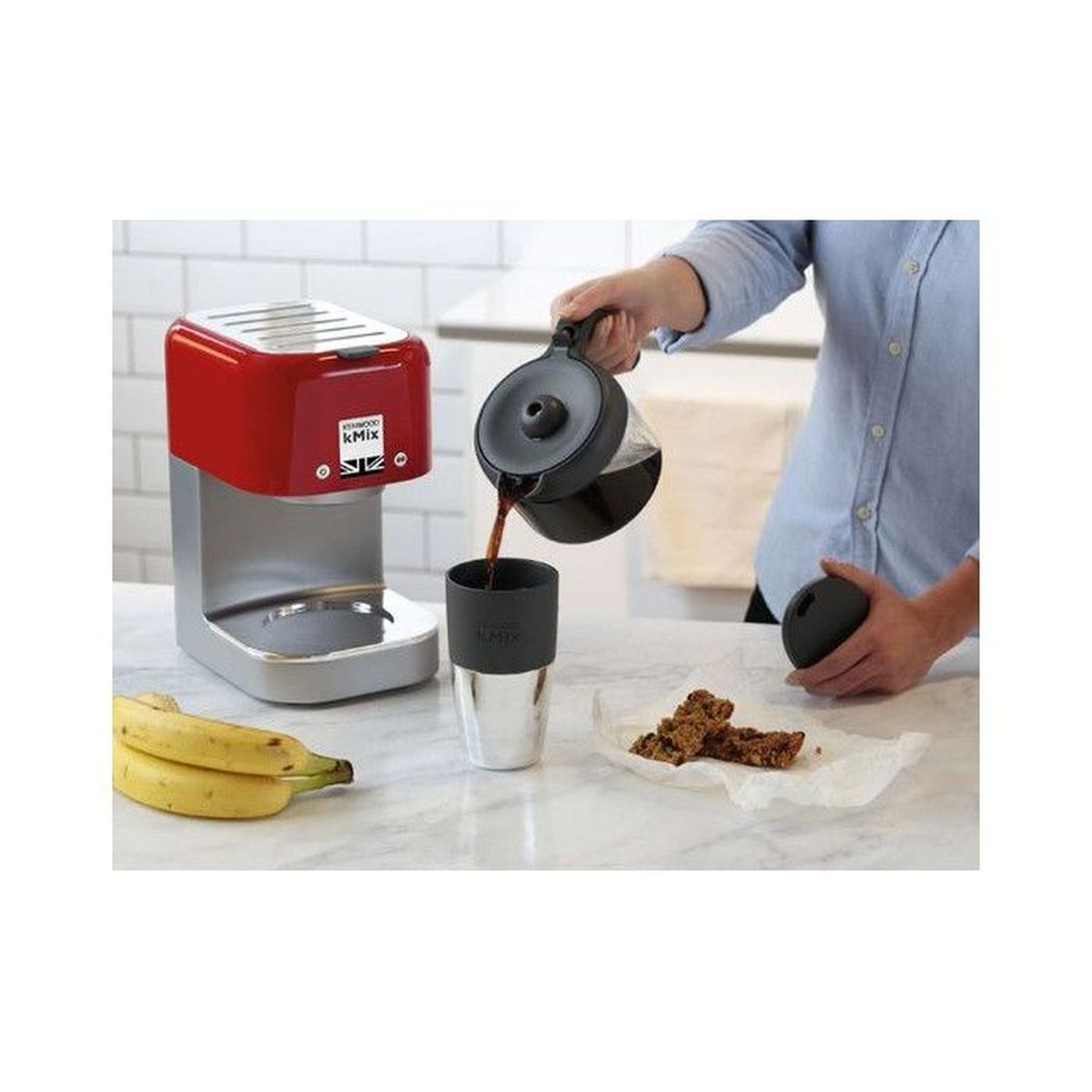 Una persona versa il caffè in una tazza termica. Banane e una macchina da caffè rossa sono sul bancone.