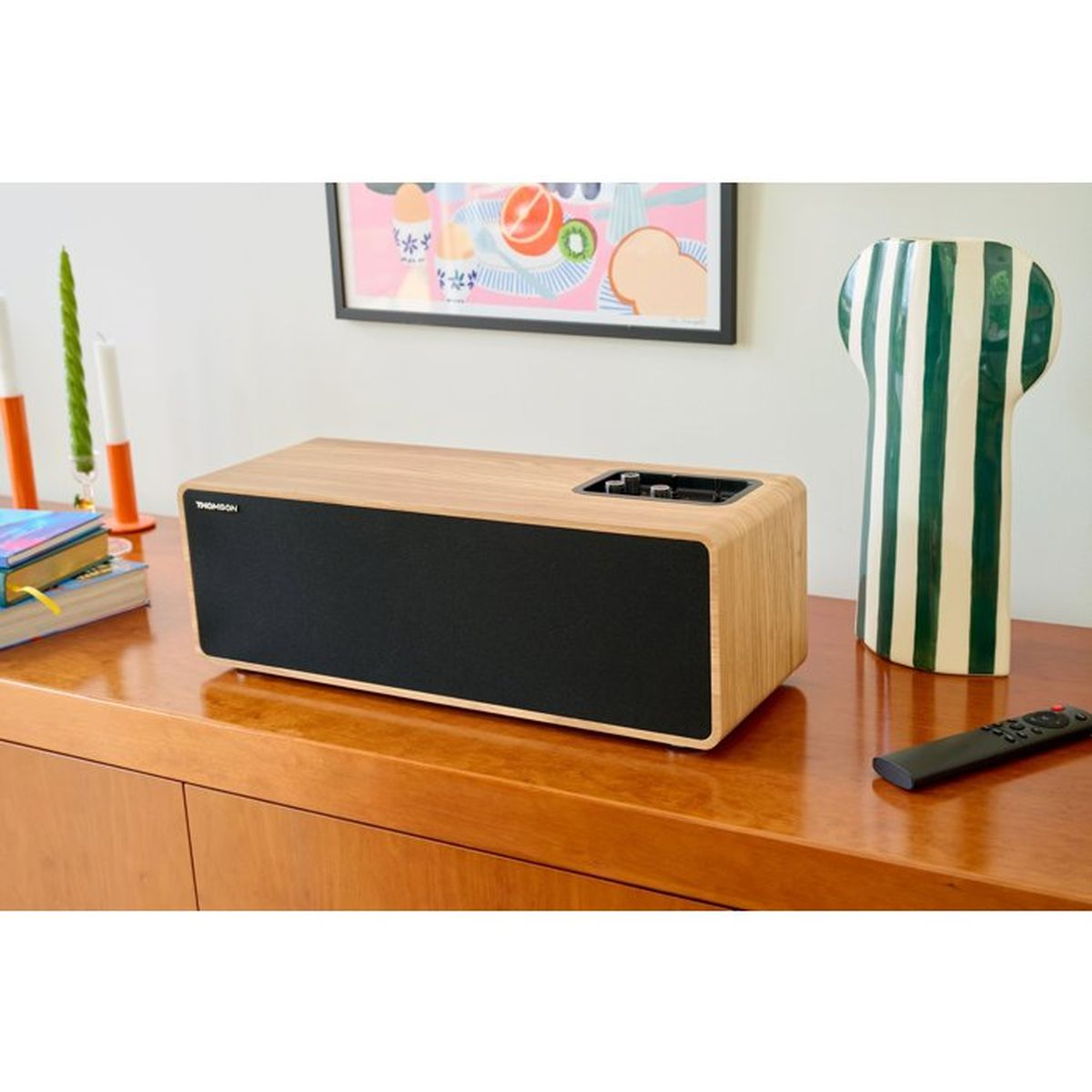 Un altoparlante in legno su una credenza con telecomando, vaso verde e bianco e quadro.