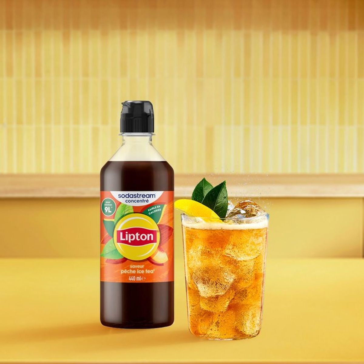 Bottiglia di concentrato di tè freddo Lipton e bicchiere con ghiaccio, limone e menta su sfondo giallo.