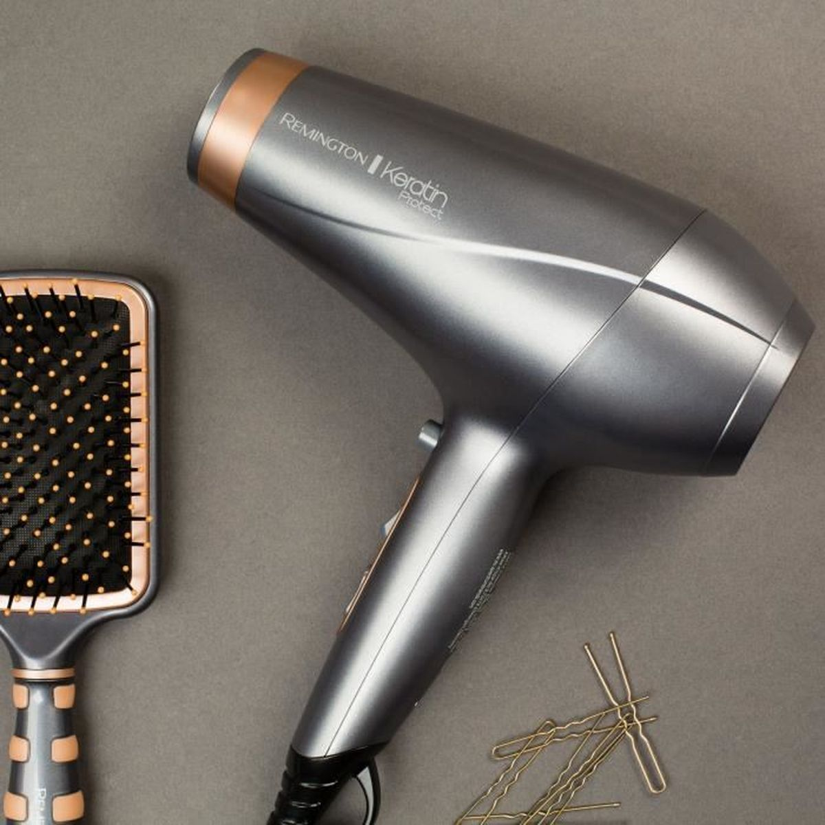 Un sèche-cheveux gris Remington Keratin Protect, une brosse et des épingles dorées reposent sur une surface grise.