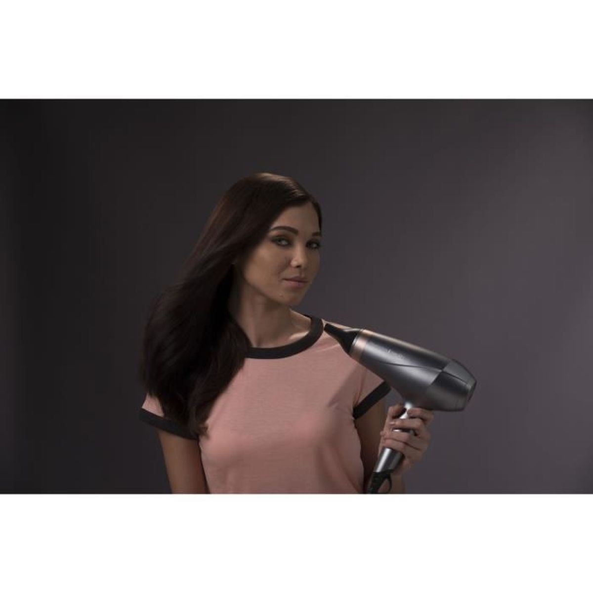 Une femme aux cheveux sombres tient un sèche-cheveux gris sur fond gris.
