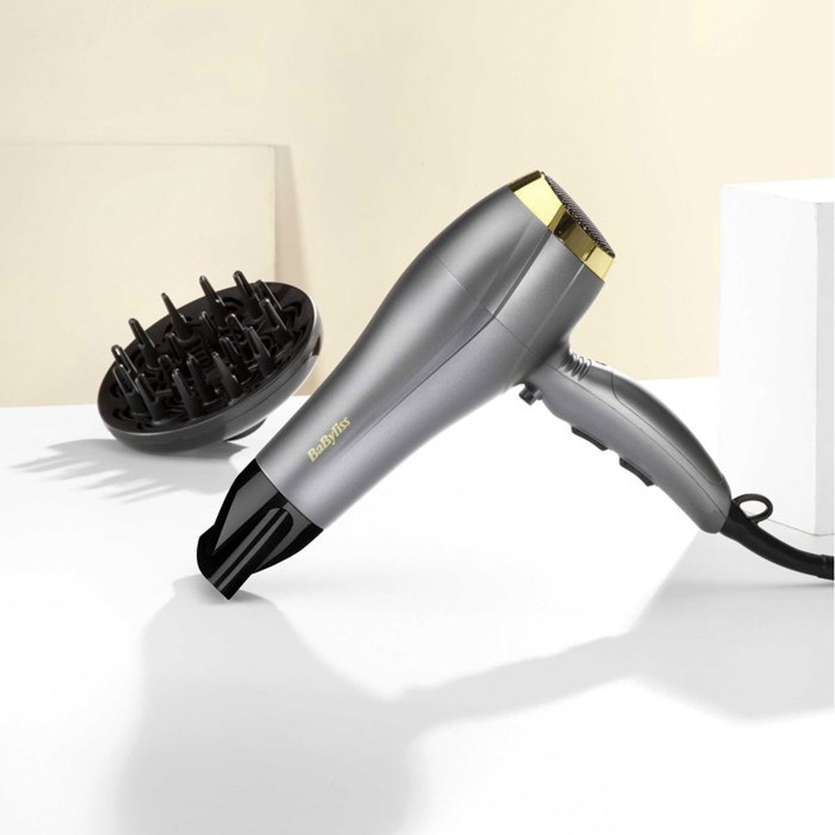 Un asciugacapelli Babyliss grigio e oro con diffusore su una superficie bianca.