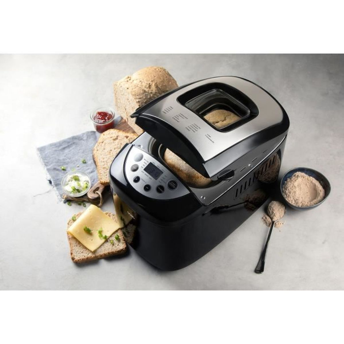 Ein schwarzer Brotbackautomat auf einer grauen Oberfläche, umgeben von Brotscheiben, Käse, Schalen mit Mehl und Aufstrich.