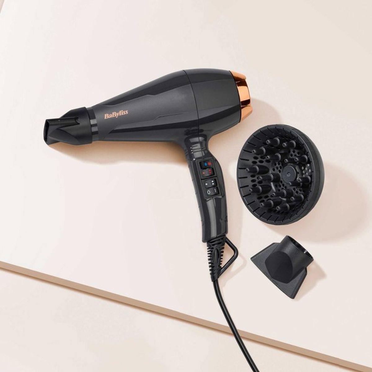 Un asciugacapelli BaByliss nero con dettagli in oro rosa, un diffusore e un beccuccio concentratore su superficie chiara.