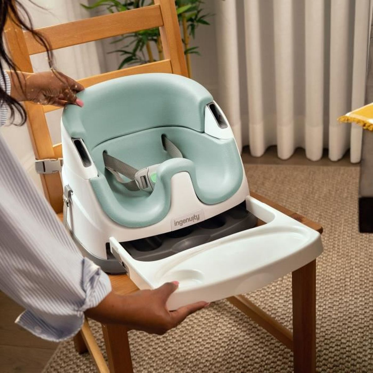 Eine Person zieht ein Tablett aus einem weiß-mintgrünen Babysitz, der auf einem Holzstuhl befestigt ist.