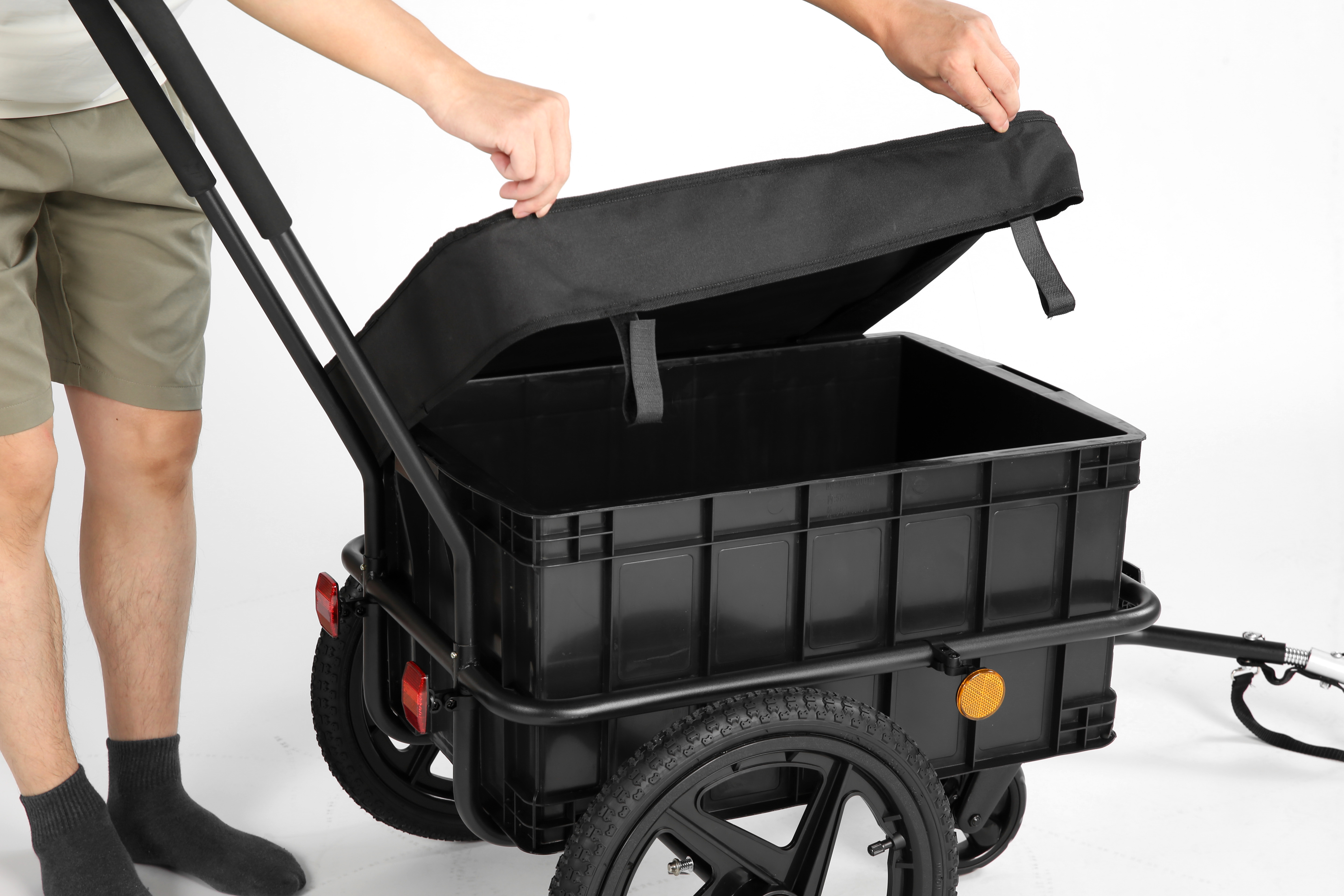 Una persona apre la copertura nera di un carrello con cassone di plastica, due ruote e sfondo bianco.