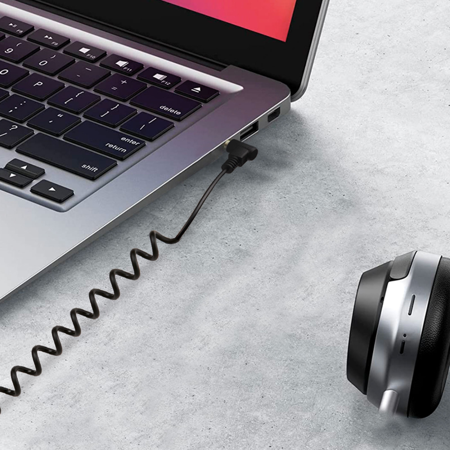 Un cable negro en espiral conecta los auriculares al puerto de audio de un portátil plateado.