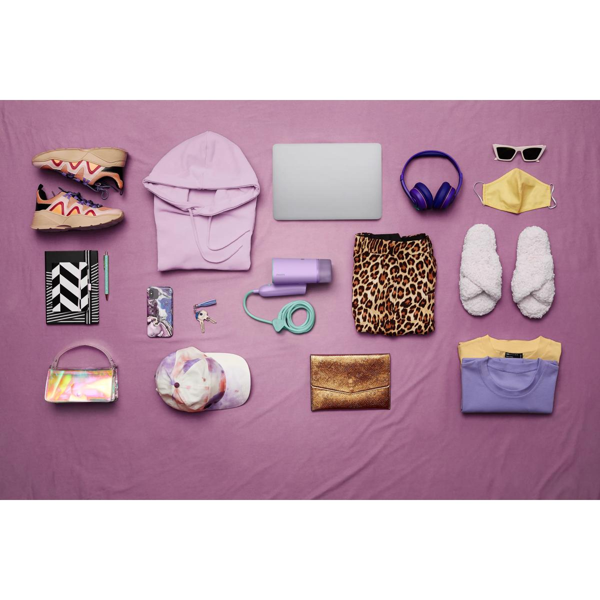 Vestiti, tecnologia e accessori disposti su superficie viola, tra cui laptop, felpa e scarpe.