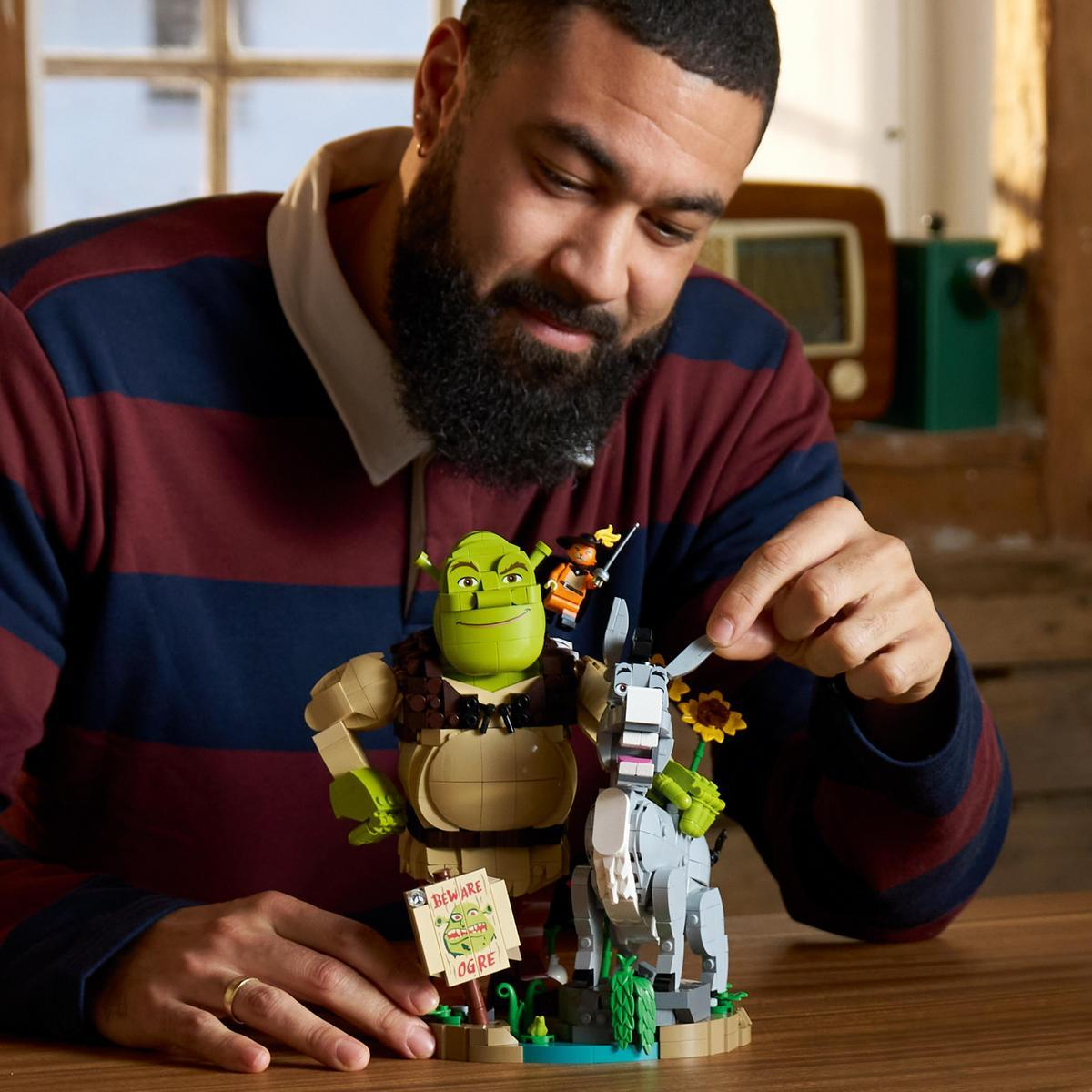Brodaty mężczyzna buduje postacie Shreka z klocków Lego na drewnianym stole.