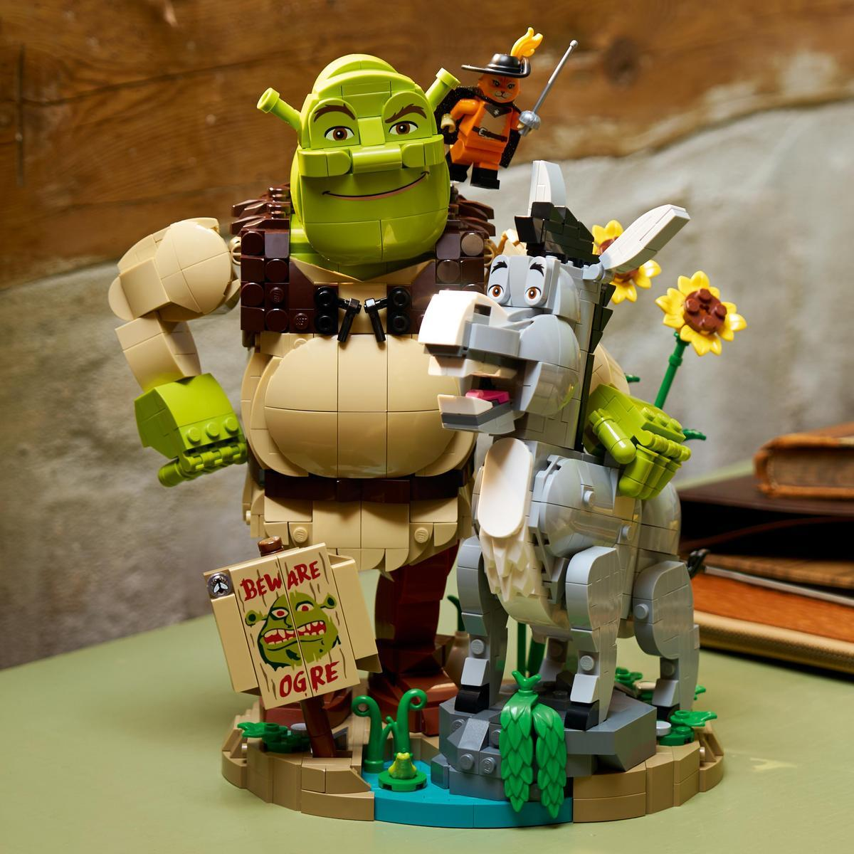 Klocki Lego przedstawiające Shreka, Osła i Kota w Butach stoją z tabliczką ostrzegającą przed ogrem na rustykalnym stole.