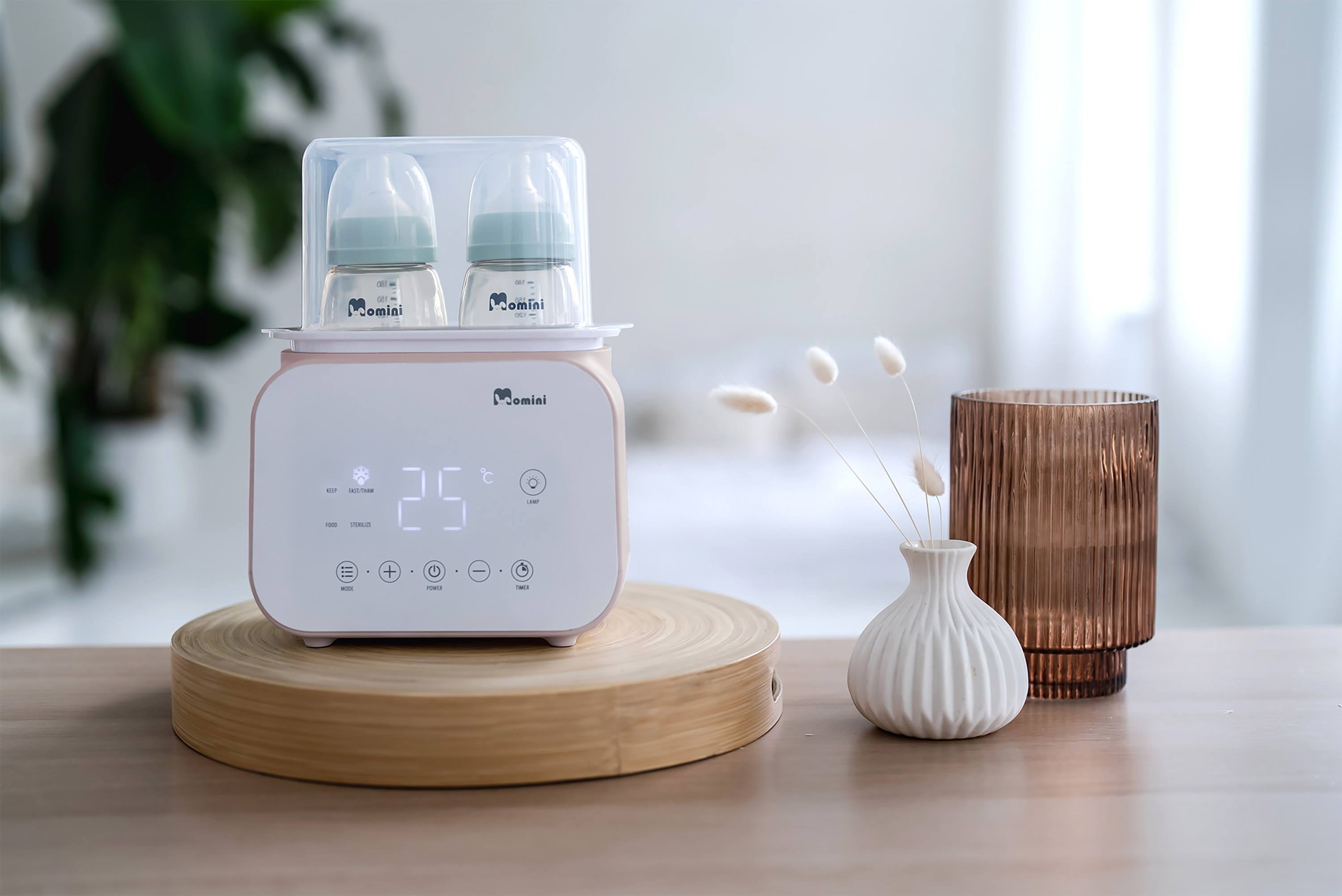 Ein rosafarbener Flaschenwärmer mit zwei Babyflaschen steht auf einem Holztisch, daneben Vasen mit Blumen.