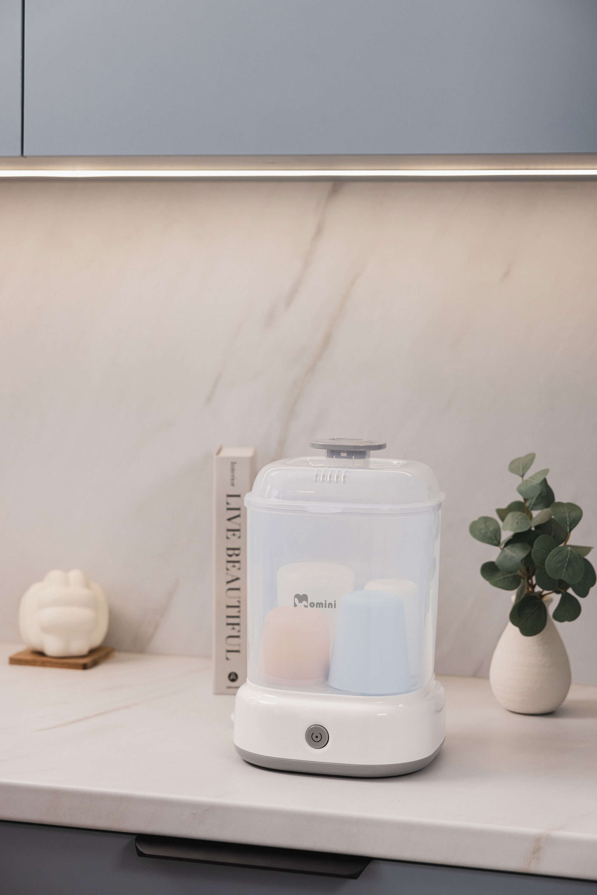 Ein weißer Babyflaschensterilisator mit bunten Flaschen steht auf einer Küchentheke mit Buch und Pflanze.