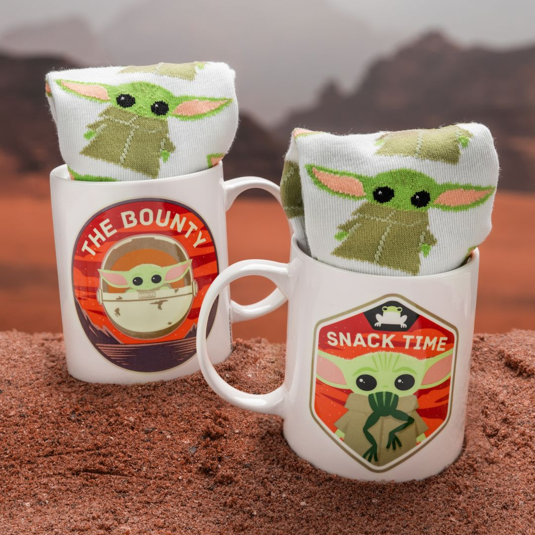 Zwei weiße Tassen mit Baby-Yoda-Grafiken und gefüllten Socken, platziert auf einer felsigen, sandigen Oberfläche.