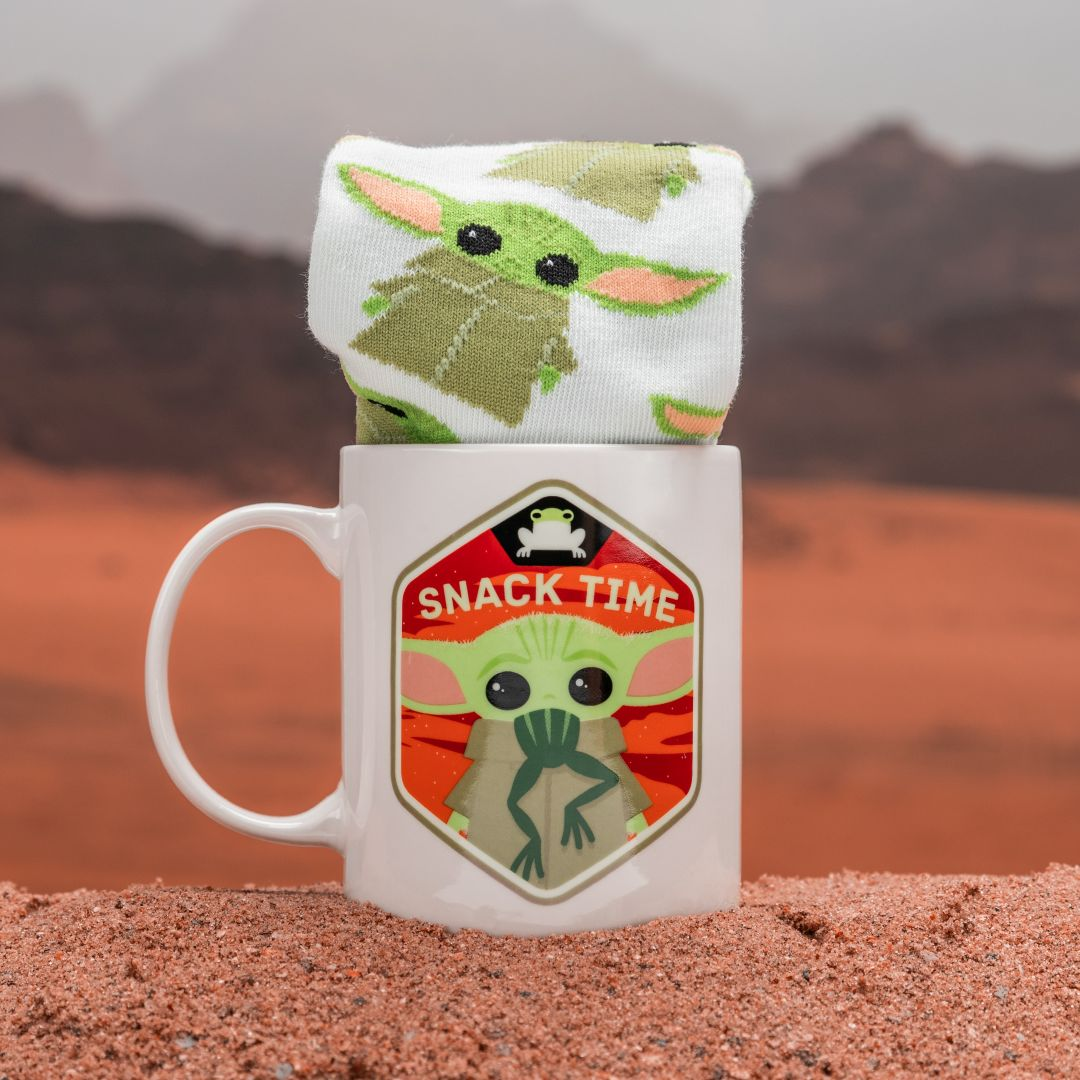 Eine weiße Tasse mit Grogu und der Aufschrift Snack Time steht im Sand, gefüllt mit Socken.