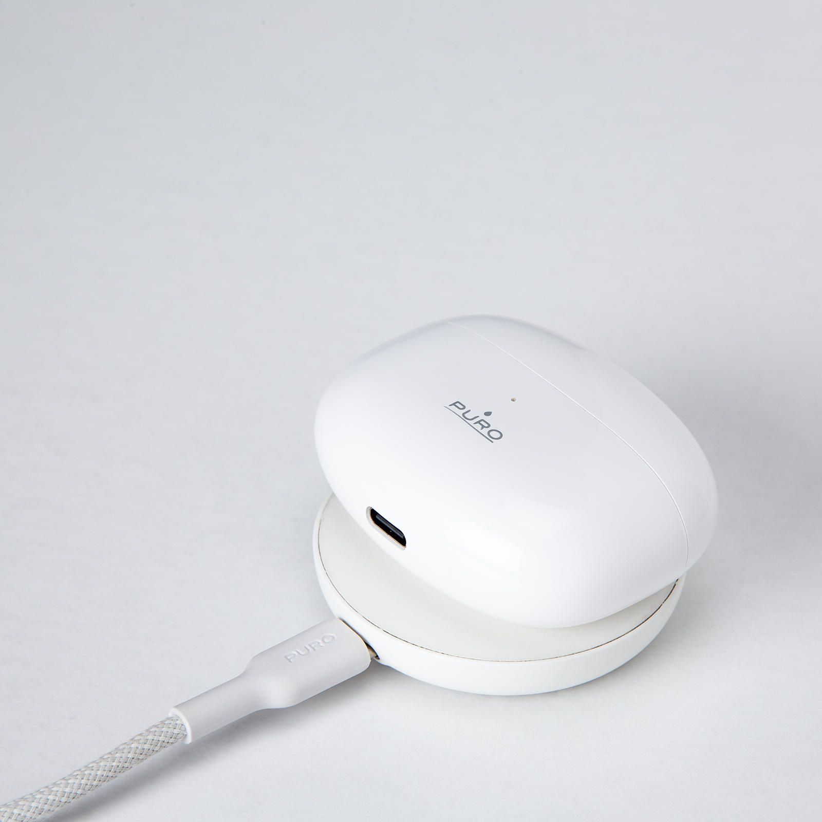 Un estuche de carga blanco de Puro descansa sobre una base de carga inalambrica con un cable sobre fondo blanco.
