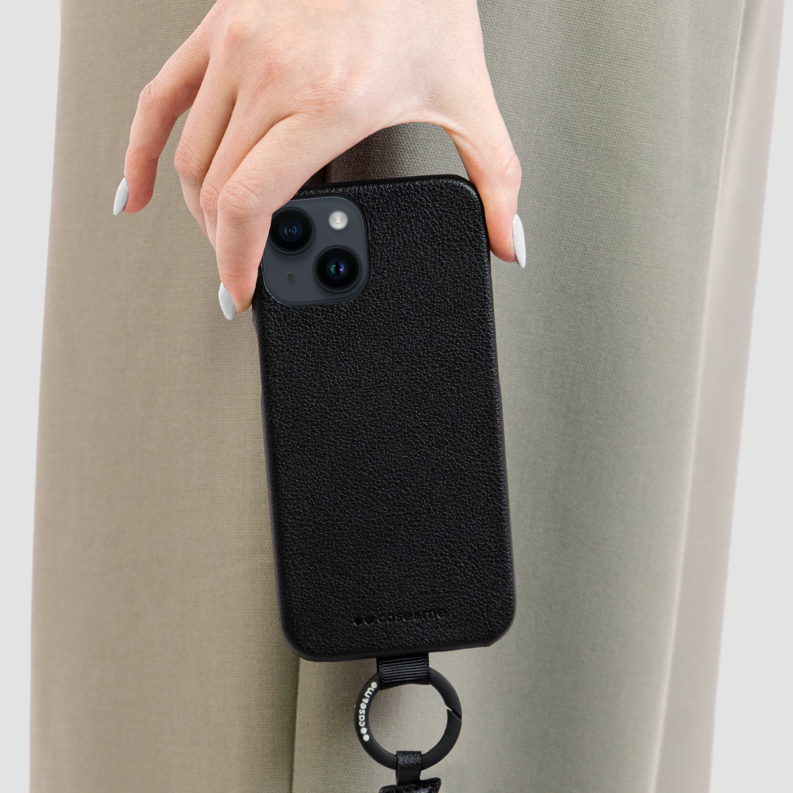 Una mano sostiene un smartphone negro con funda texturizada y correa negra contra pantalones grises.