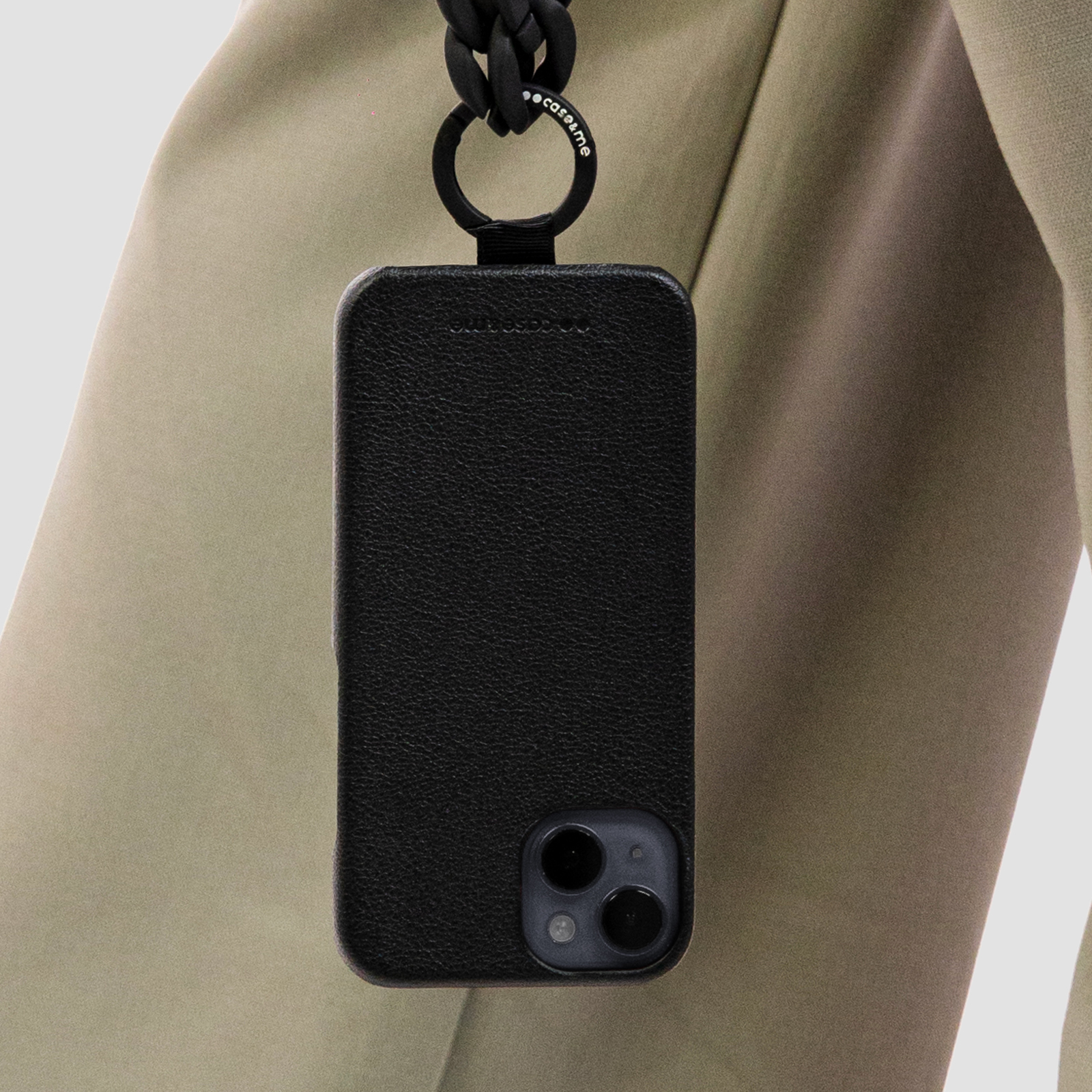 Una funda de móvil de cuero negro con correa, colgada sobre fondo de prenda beige.