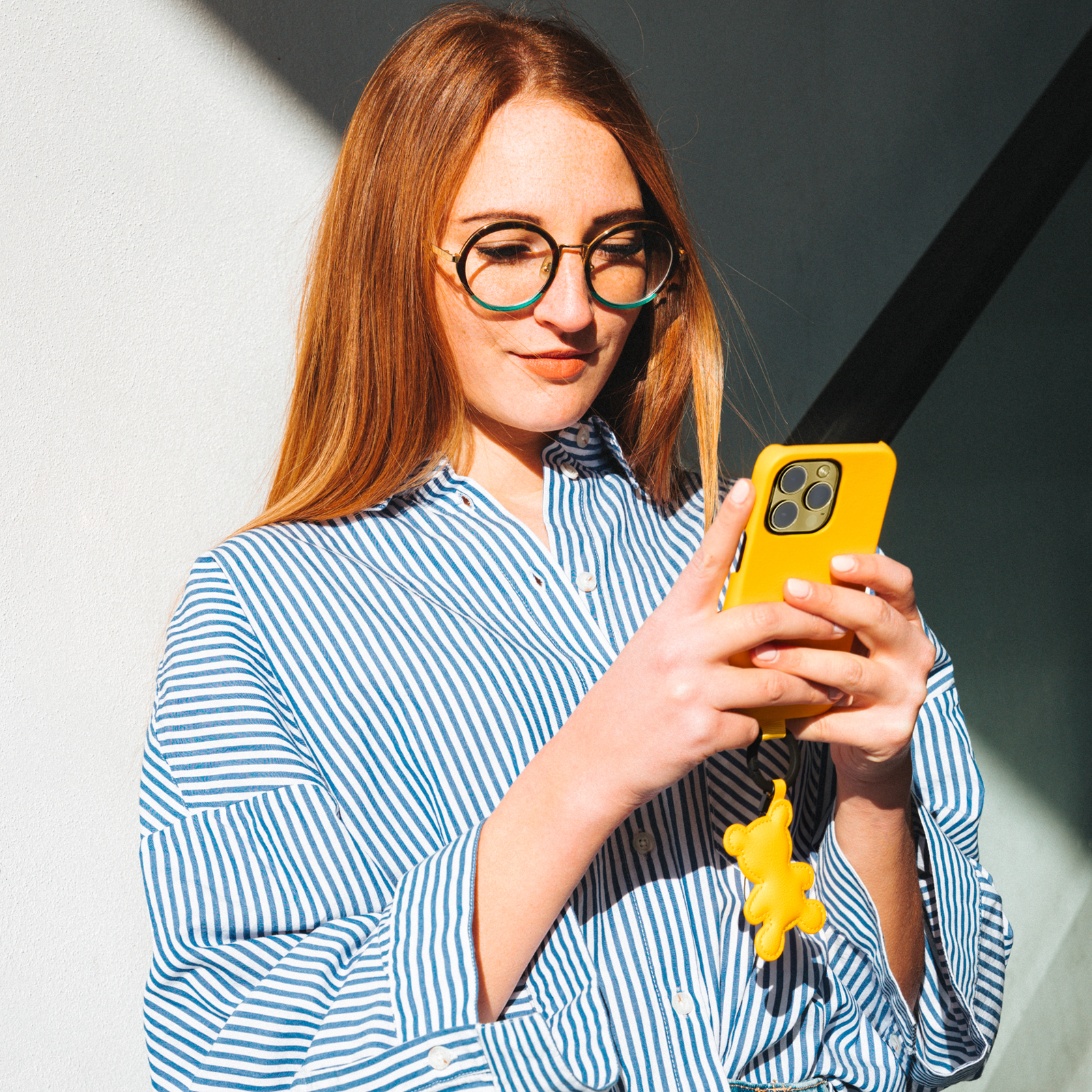 Una mujer pelirroja con gafas y camisa de rayas azul y blanca escribe en su smartphone amarillo.