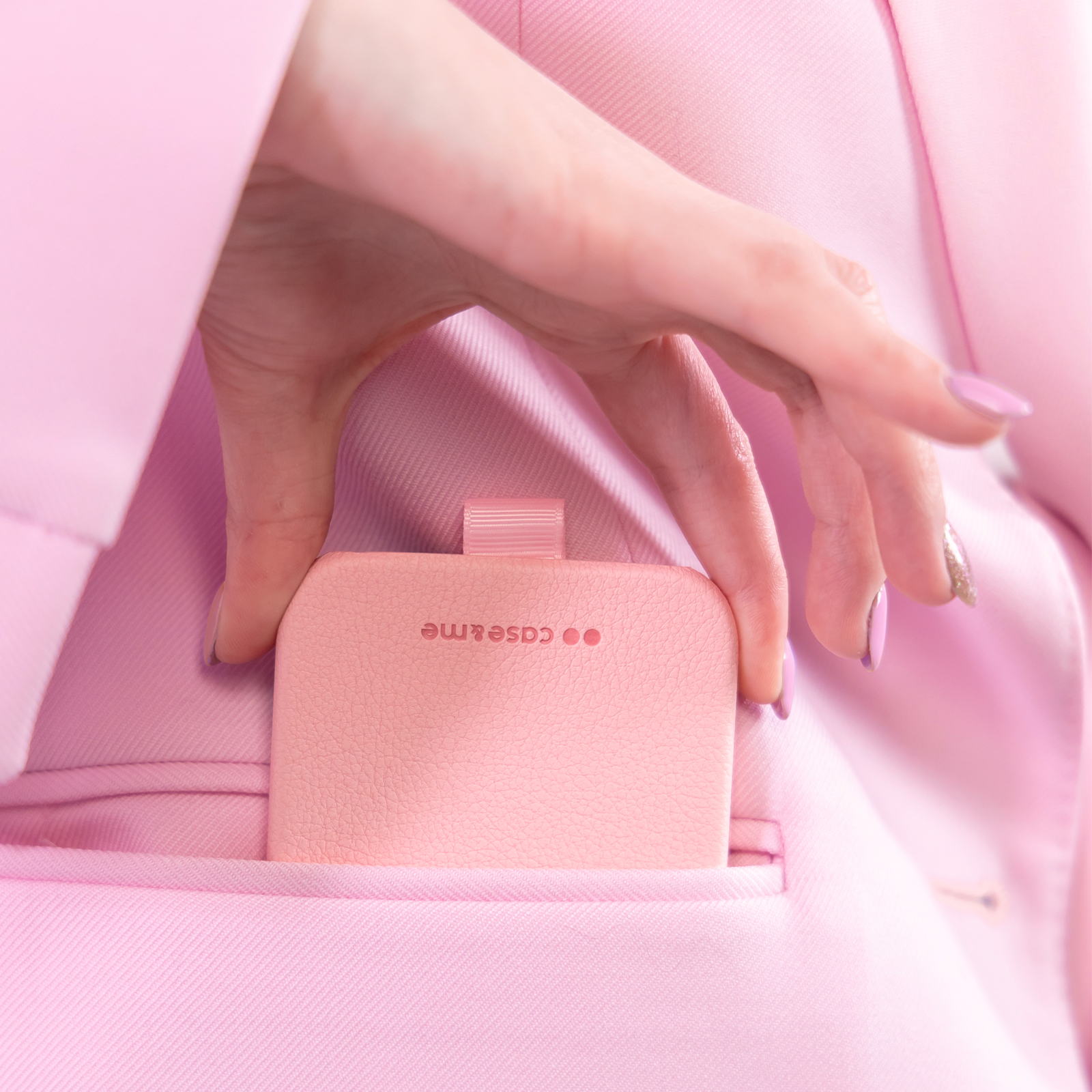 Una mano coloca un estuche de cuero rosa con el texto "case&me" en el bolsillo de un blazer rosa.
