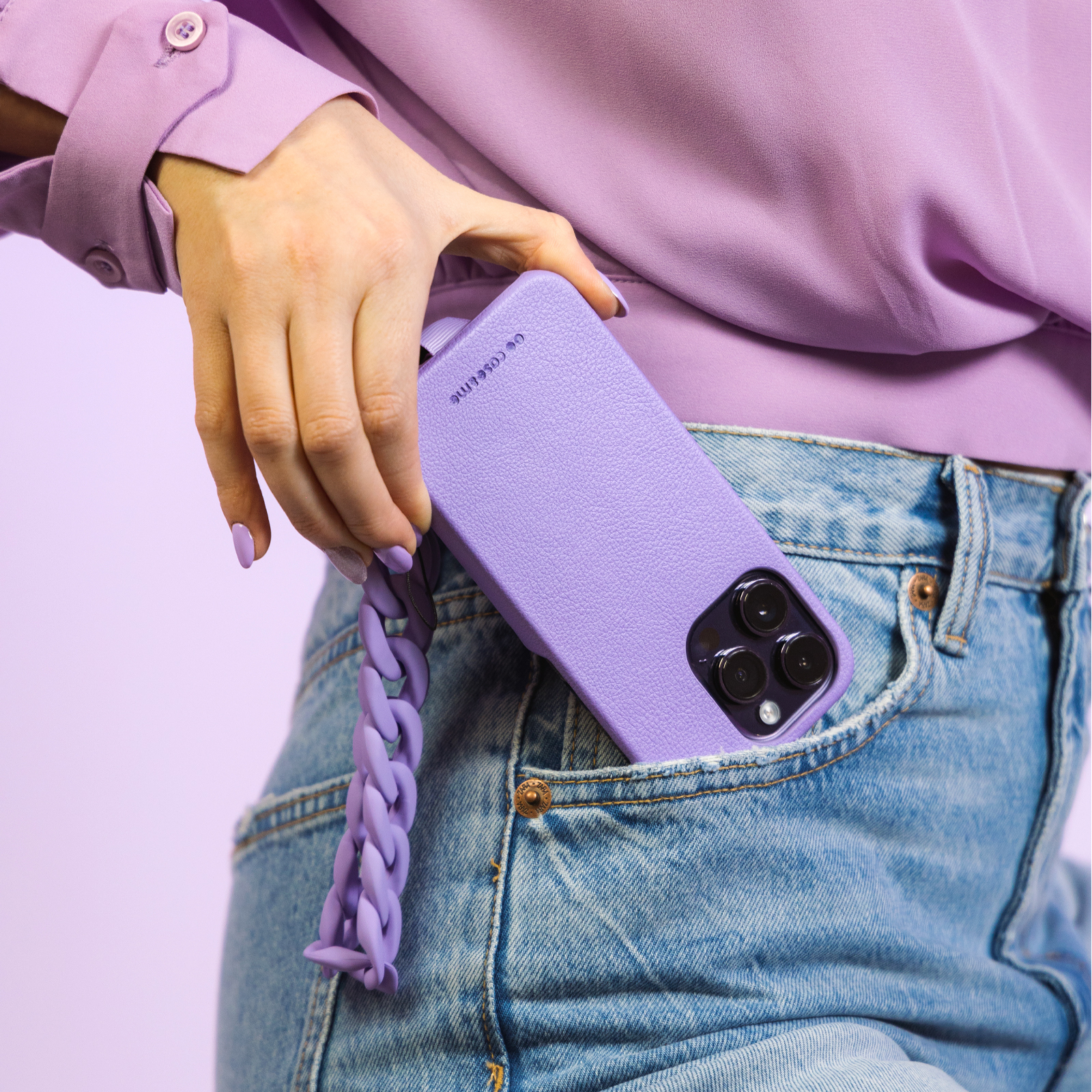 Mano che tiene un telefono viola con catena nella tasca dei jeans, su sfondo rosa.