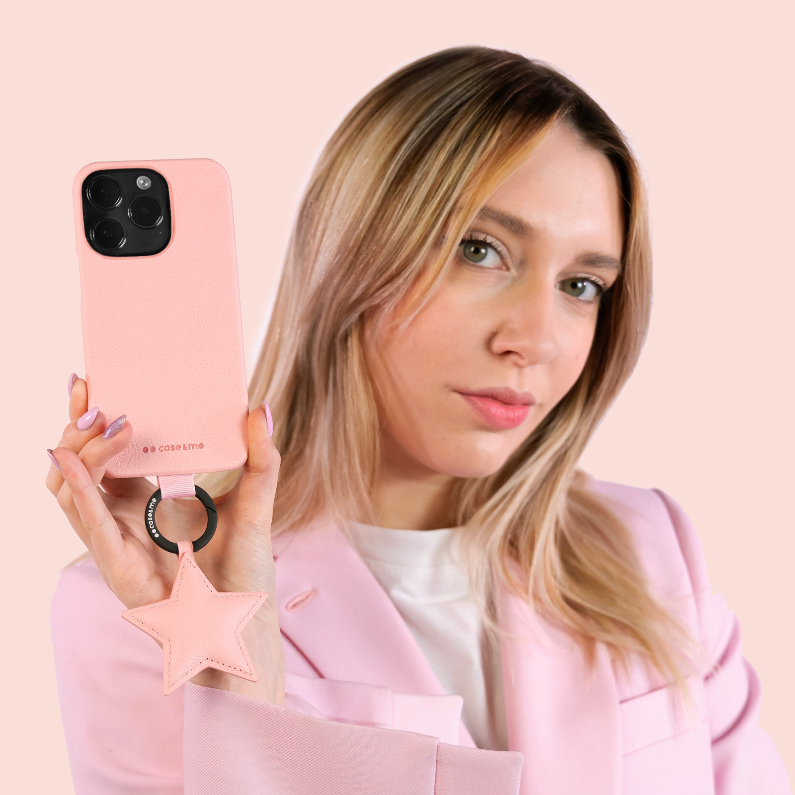 Una mujer rubia con chaqueta rosa sostiene un móvil rosa con un colgante de estrella ante un fondo liso.