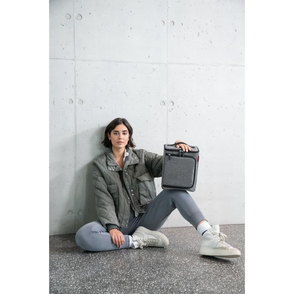 Une femme en veste et legging gris est assise contre un mur en béton, tenant un sac isotherme gris.