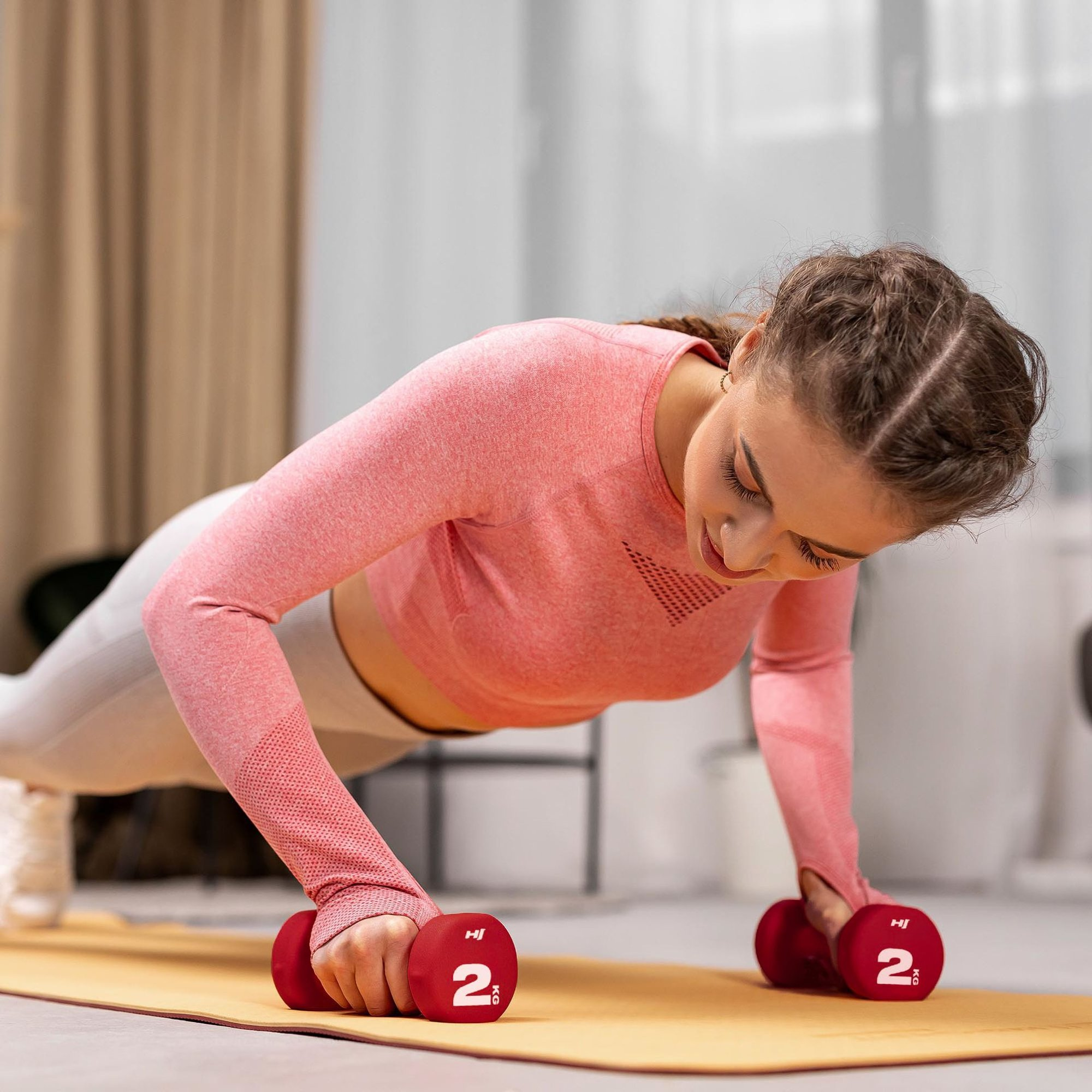 Eine Frau in Sportkleidung macht Liegestütze auf einer gelben Matte und hält zwei rote Hanteln.