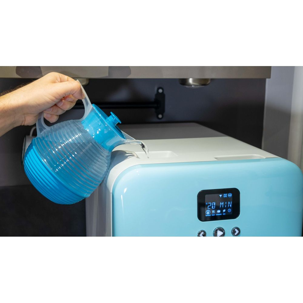 Una mano versa acqua da una caraffa blu nel serbatoio di un apparecchio azzurro con display.