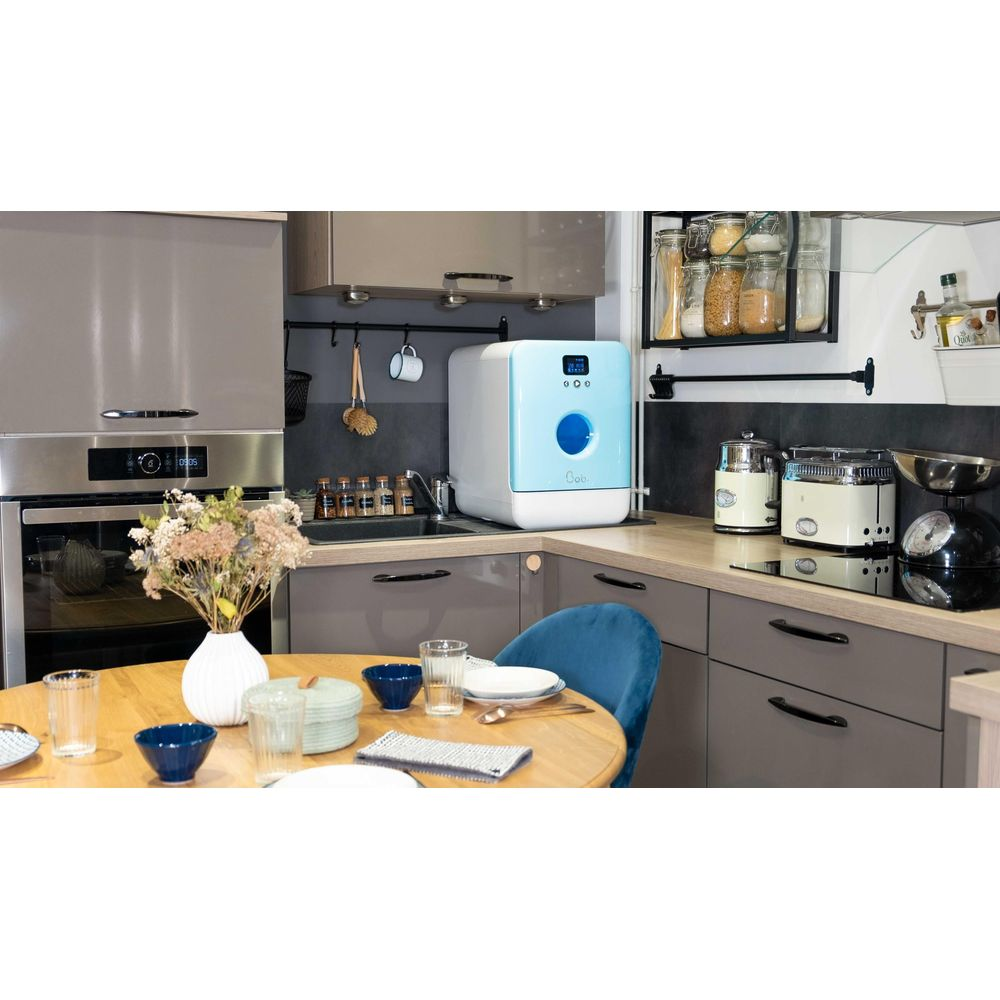 Moderna cucina con mobili grigi, tavolo in legno con fiori e una lavastoviglie azzurra sul piano di lavoro.