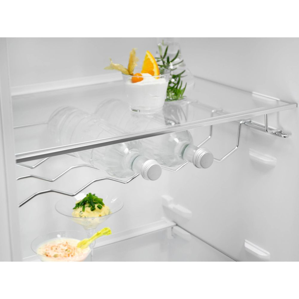All'interno di un frigorifero bianco, una griglia regge due bottiglie, con un dessert e erbe aromatiche sopra.