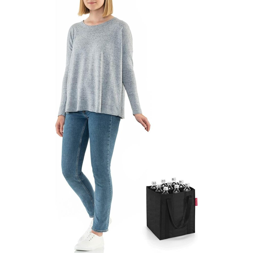 Eine Frau in grauem Pullover und Jeans steht neben einer schwarzen Einkaufstasche mit mehreren Flaschen.