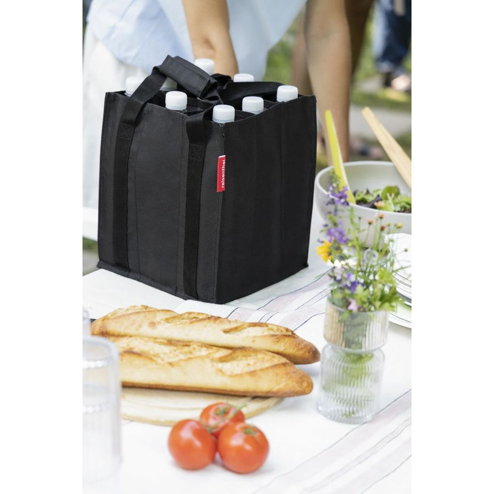 Eine schwarze Flaschentasche steht auf einem Picknicktisch mit Baguettes, Tomaten und einer Blumenvase.