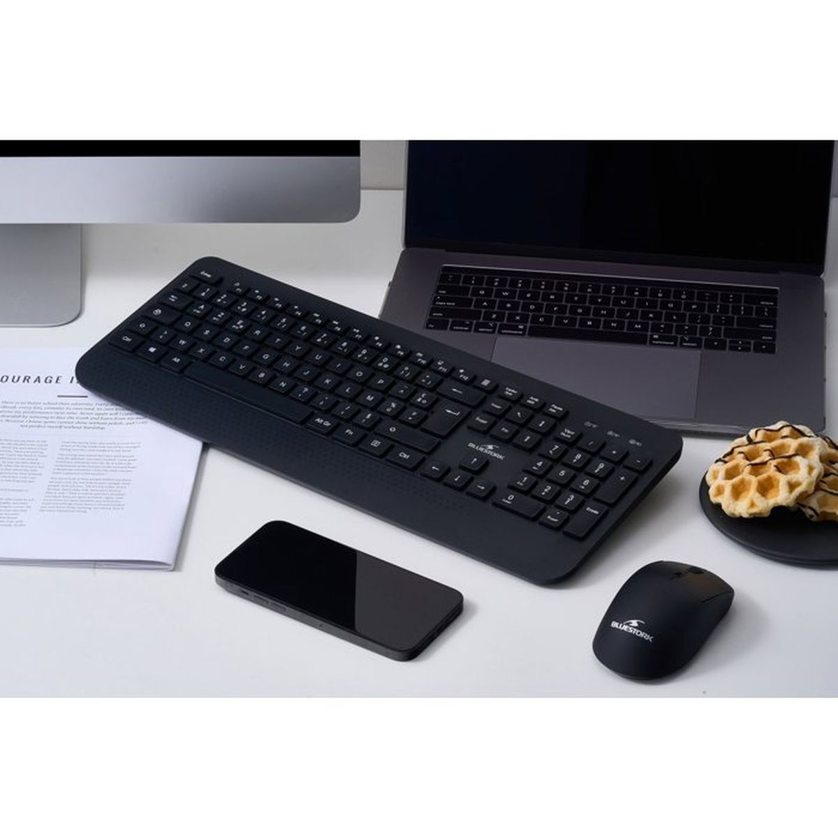 Una tastiera, un mouse e uno smartphone neri su una scrivania bianca con laptop.