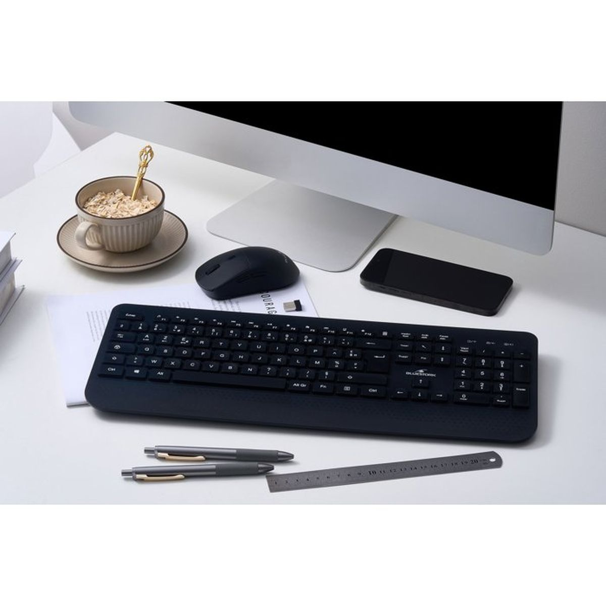 Una scrivania bianca con tastiera nera, mouse, smartphone, monitor, tazza di cereali, due penne e un righello.