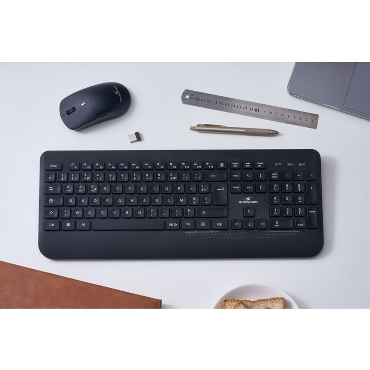 Una scrivania con tastiera wireless nera, mouse, ricevitore USB, righello metallico, penna, laptop, taccuino e toast.