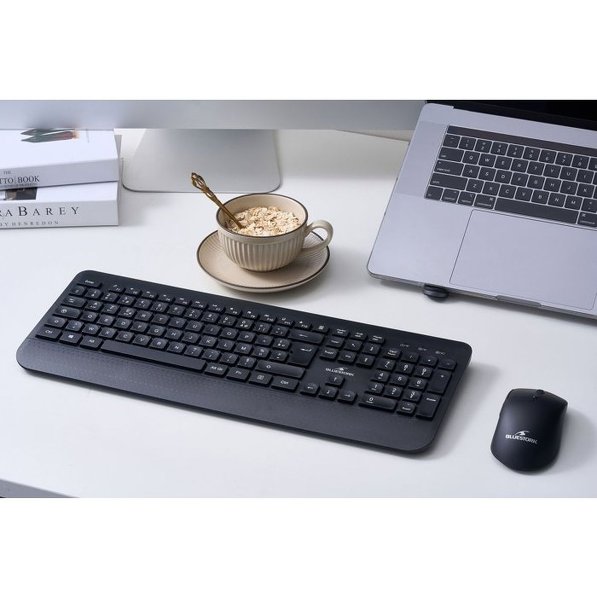 Una scrivania con laptop, tastiera nera, mouse, libri e una tazza di cereali con cucchiaio.