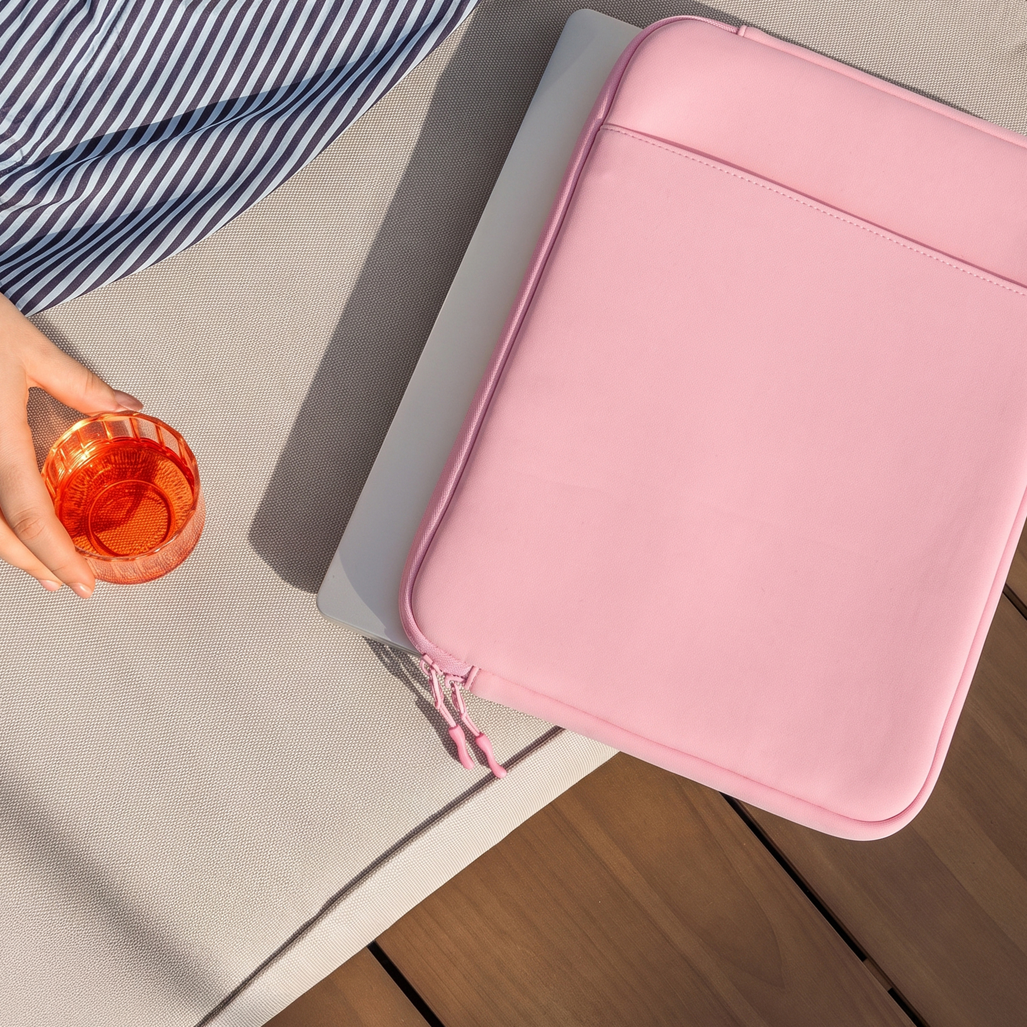 Eine Hand hält ein Glas mit einem orangefarbenen Getränk neben einer pinken Laptoptasche auf einem Liegestuhl.