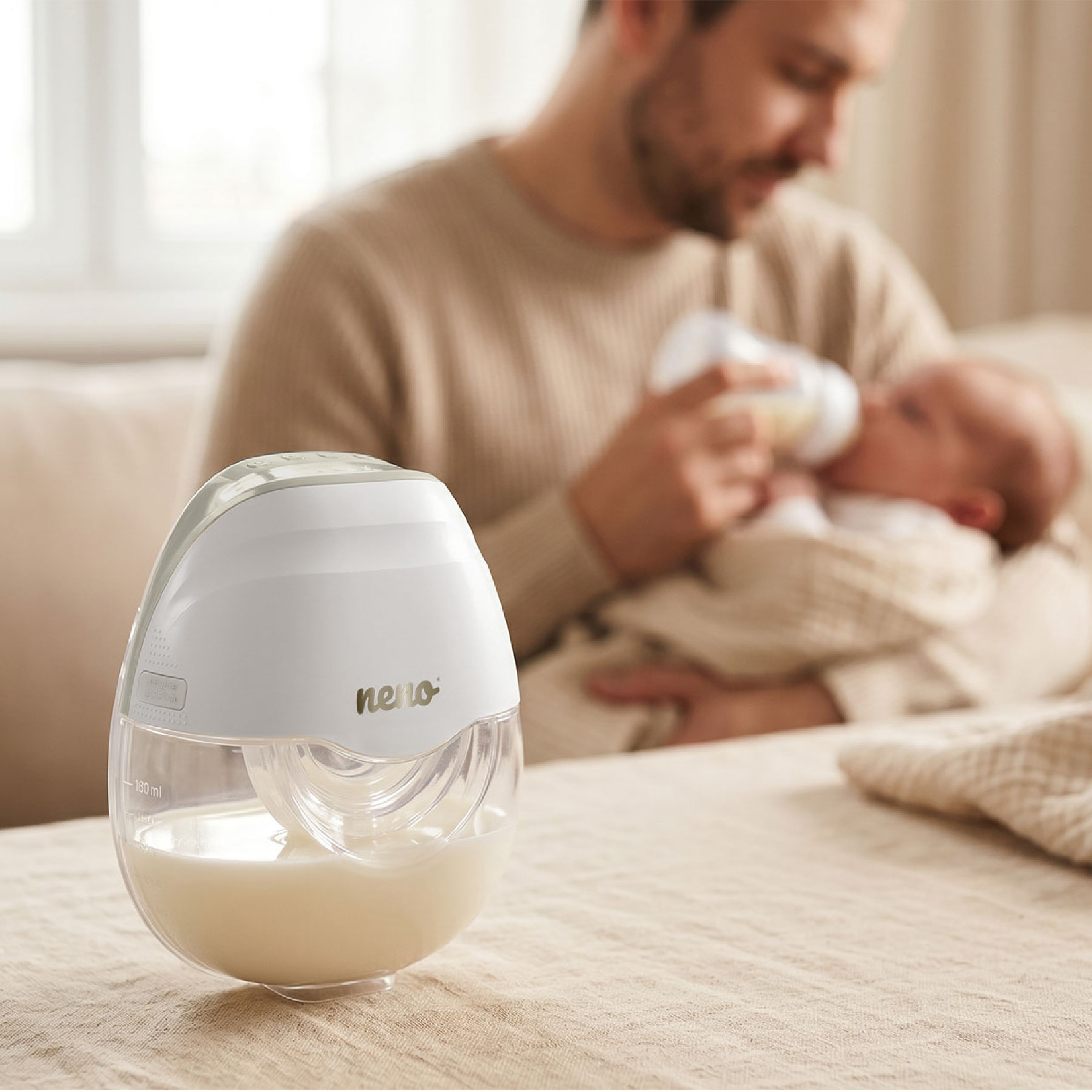 Eine weiße Milchpumpe steht im Fokus auf einem Tisch, während im Hintergrund ein Vater sein Baby mit der Flasche füttert.
