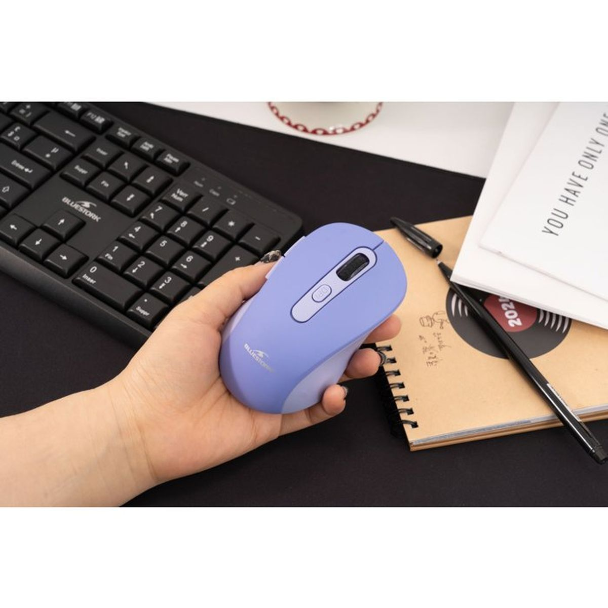 Una mano sostiene un ratón de ordenador lavanda sobre un escritorio con teclado y cuaderno.