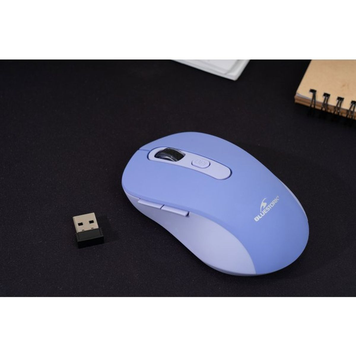 Un ratón inalámbrico azul claro con un receptor USB en una superficie negra, junto a un cuaderno.